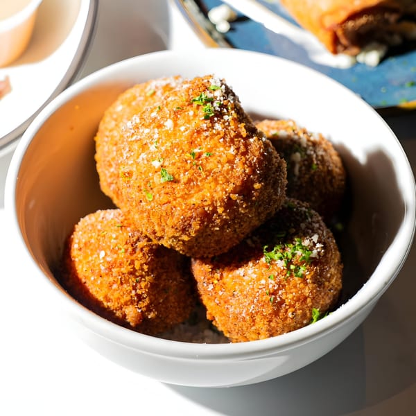 Close-up of golden-brown arancini balls, sprinkled with cheese and parsley, in a white bowl.