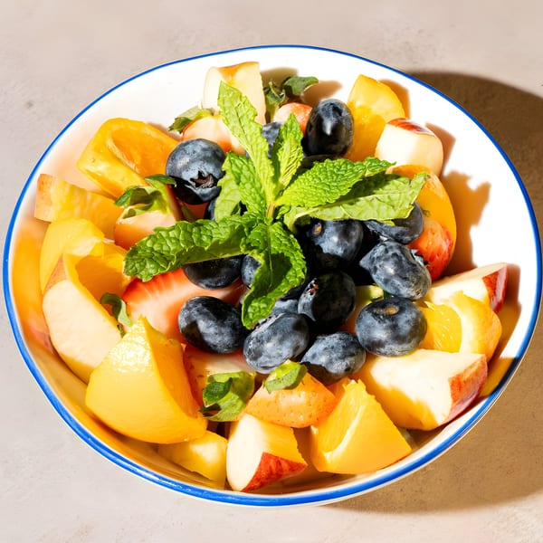 A colorful fruit salad with blueberries, strawberries, peaches, and mint in a white bowl.