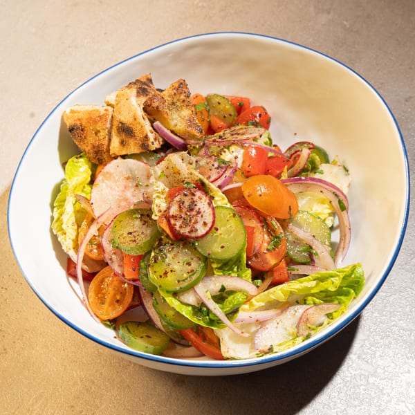 A fresh salad with lettuce, tomatoes, cucumbers, red onions, and grilled bread in a white bowl.