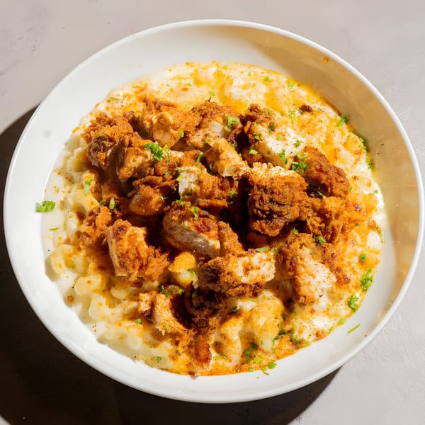 A white bowl filled with creamy macaroni and cheese topped with crispy fried chicken pieces.