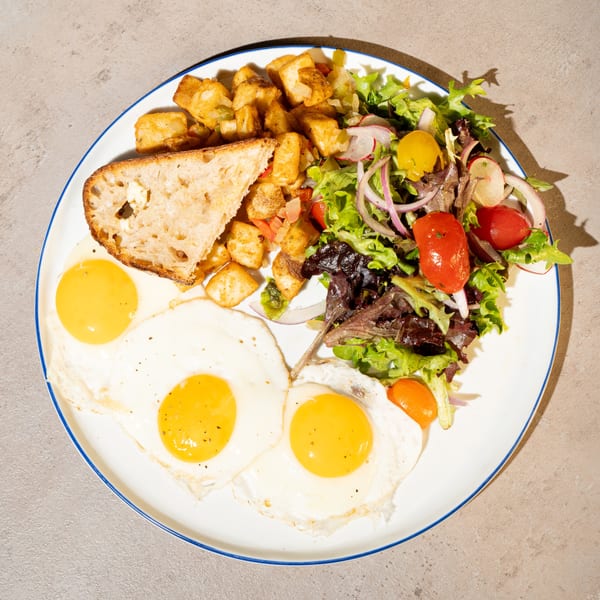 A breakfast plate with three fried eggs, home fries, a side salad, and a slice of toast.
