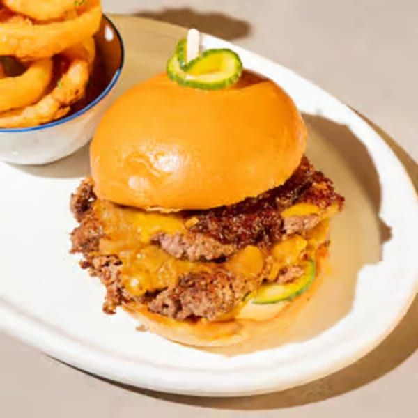 Double cheeseburger with pickles and a side of onion rings on a white plate.